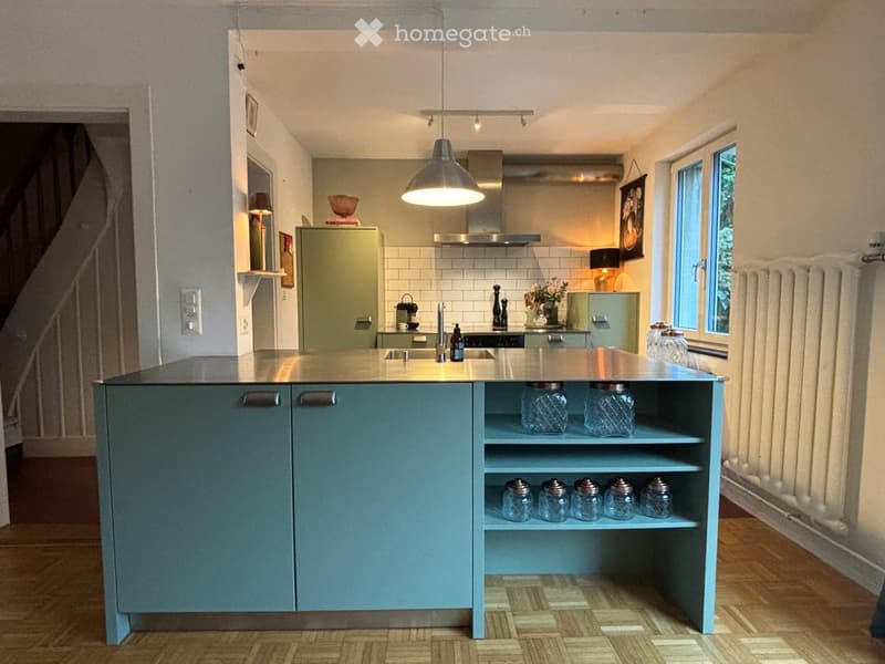 Modern kitchen with a central island, stainless steel sink, and various appliances. The island features open shelving with glass jars and a pendant light hangs above.