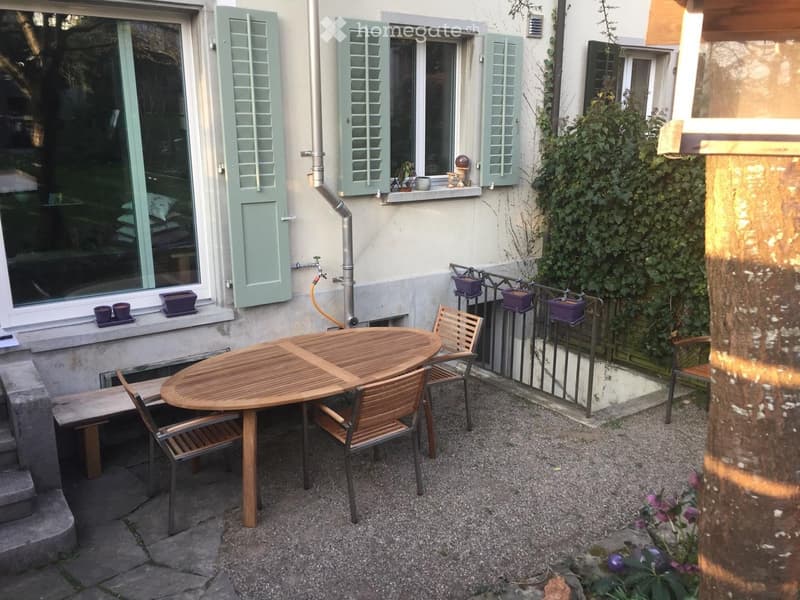 Small outdoor patio with a round wooden table, four matching chairs, a bench, and several potted plants on the windowsills.