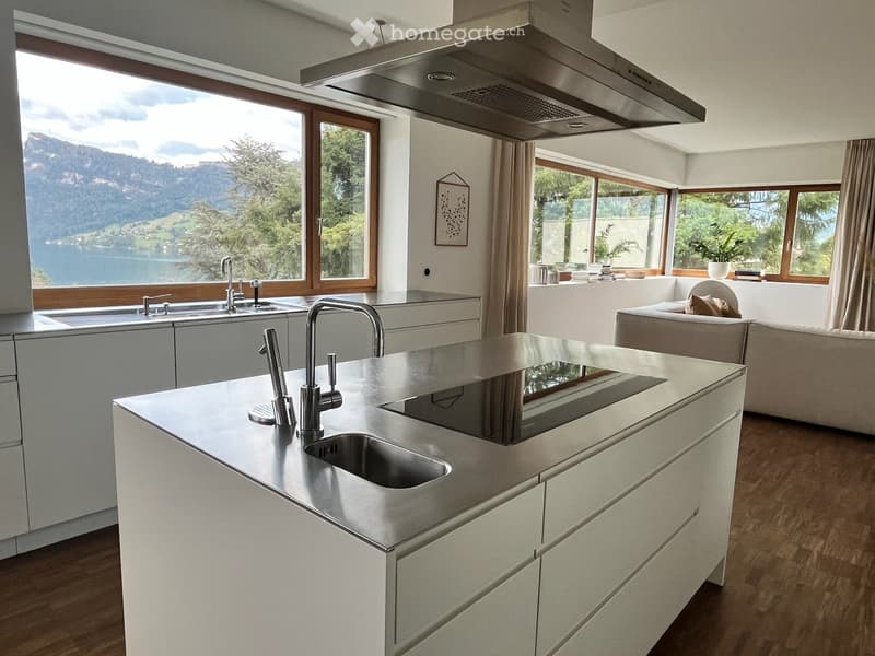 Modern kitchen with white cabinets, stainless steel island, double sink, and large windows offering a scenic lake view. A small seating area with a sofa is visible in the background.