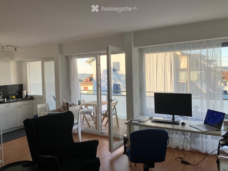 A modern, well-lit room with a desk setup, a dining area, and a balcony view. The room features a large window with sheer curtains, a sliding glass door leading to a balcony, and a mix of contemporary furniture including a black armchair, a wooden dining