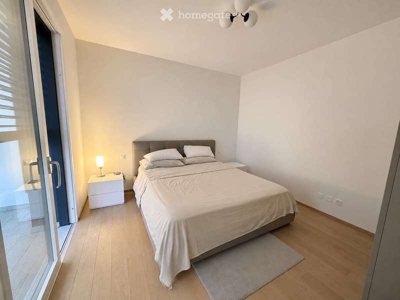 Modern bedroom with a large bed, nightstand, lamp, and a rug on a wooden floor. The room features white walls, a large window with blinds, and a sleek, contemporary design.