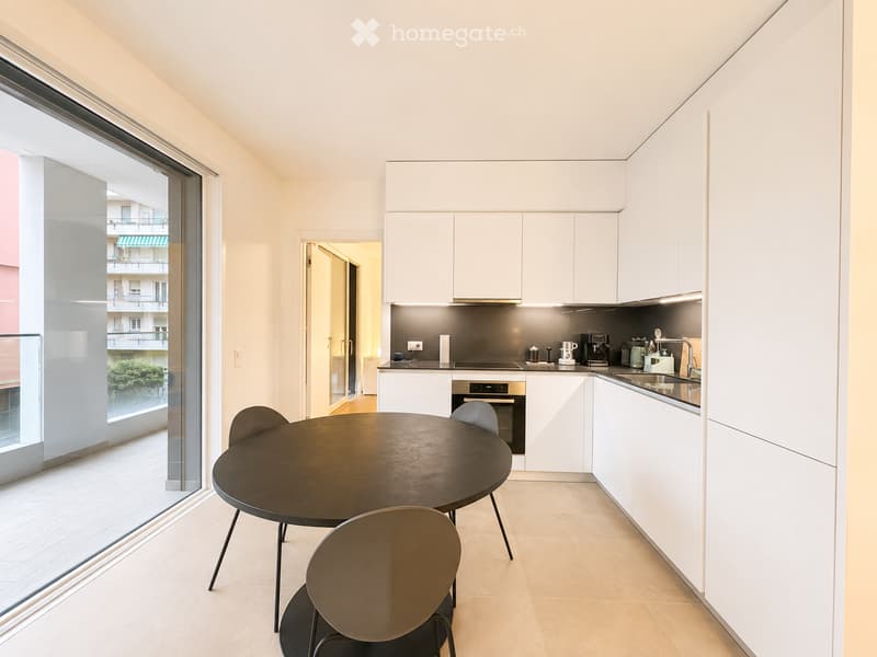 Modern kitchen with white cabinets, stainless steel appliances, a round dining table with chairs, and a large window offering a view of the building exterior.