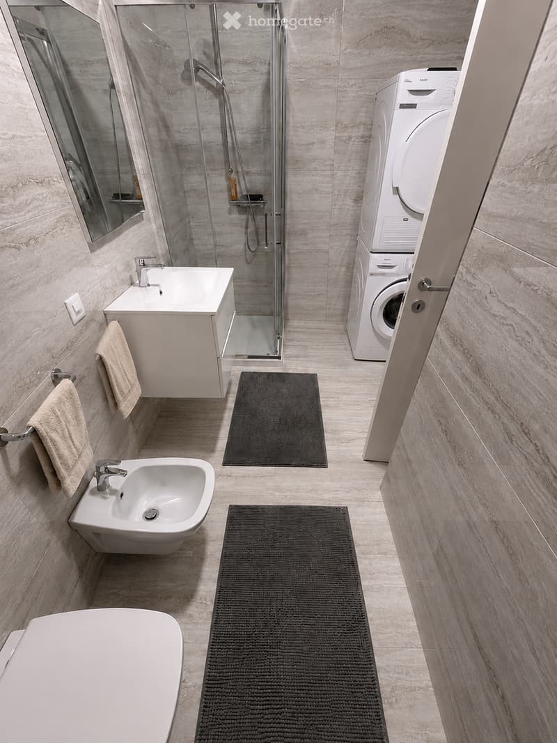 Modern bathroom with marble walls and floors, featuring a bathtub, sink, mirror, shower, washing machine, and towels.