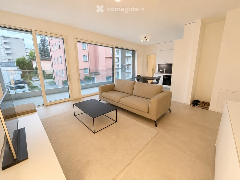 Modern living room with large sliding glass doors leading to a balcony, beige sofa, black coffee table, dining area with chairs and table, and a small kitchen area with an oven and screen.