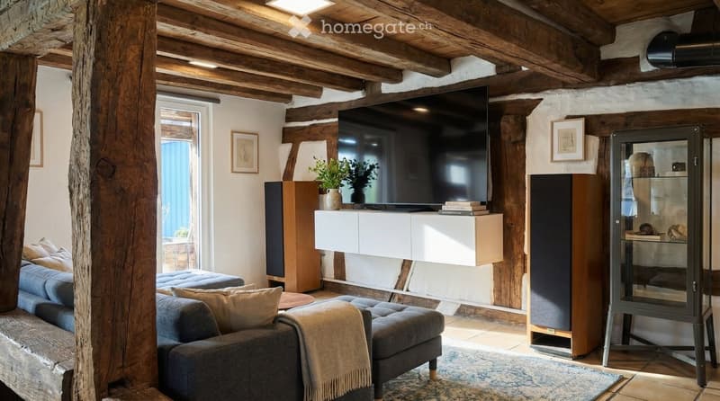 Living room with wooden beams, gray couch, ottoman, rug, TV, speakers, cabinet, plants