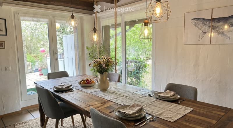Furnished Room. Dining area with a wooden table, chairs, and a floral centerpiece. The table is set with plates, napkins, and cutlery. The room has large glass doors leading to a garden.