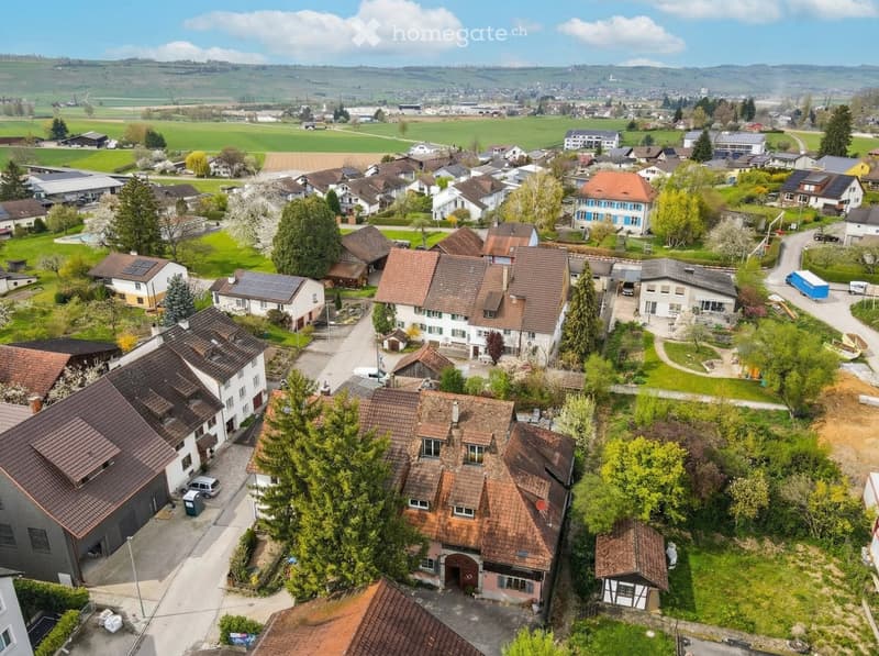 Aerial view of a neighborhood with several houses, some with solar panels, surrounded by green fields and trees.