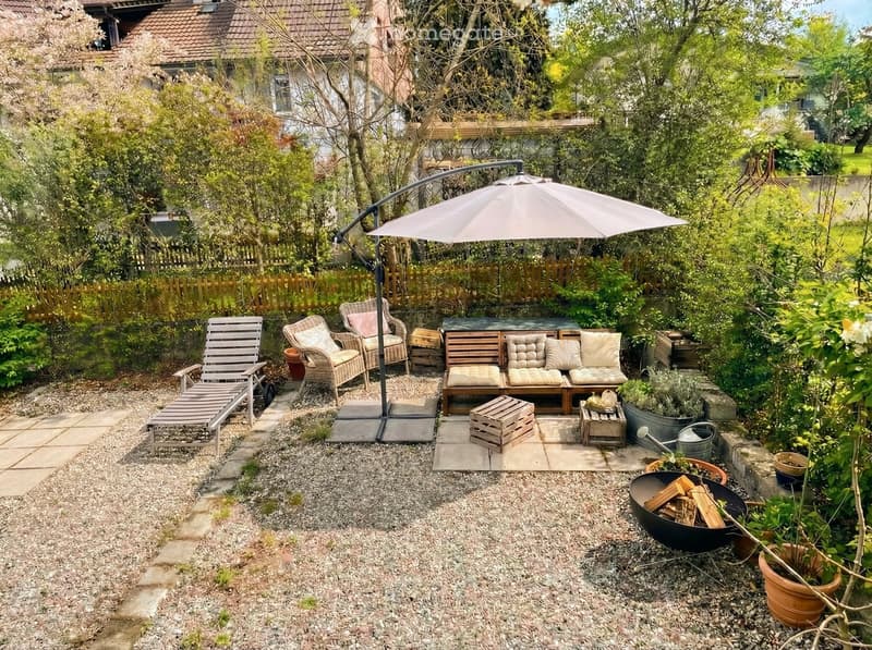 Outdoor seating area with a lounge chair, a wooden bench, a wooden table, and a large umbrella. Potted plants and shrubs are scattered around the area. A wooden fence surrounds the area, and a house is visible in the background.