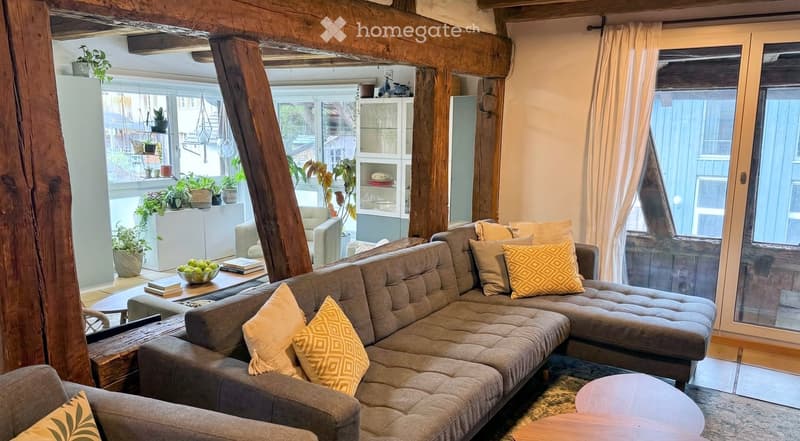 L-shaped sofa, coffee table, armchair, wooden beams, glass doors, balcony, plants, books, bowl of fruit