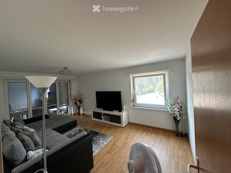 Modern living room with wooden flooring, large windows, grey sectional sofa, coffee table, TV stand, floor lamp, fan, and decorative vases.