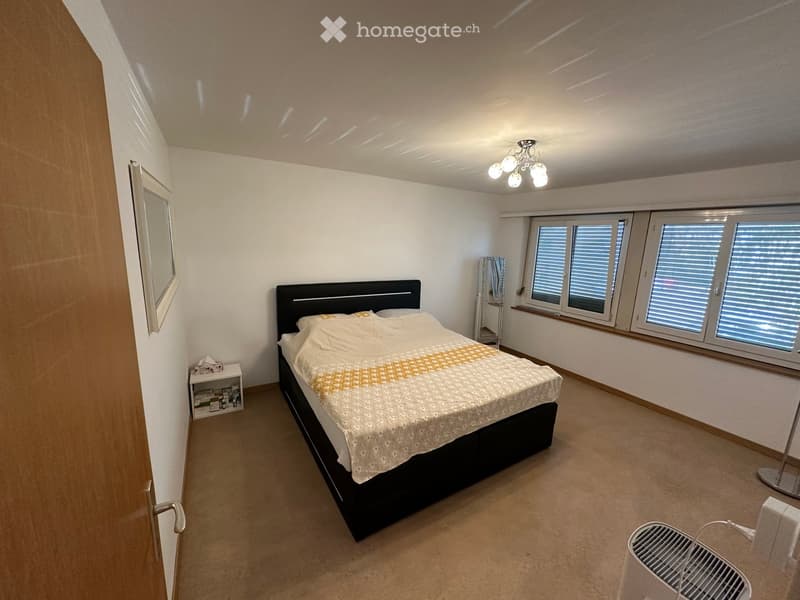 Modern bedroom with a large bed, nightstand, mirror, and large windows with blinds.