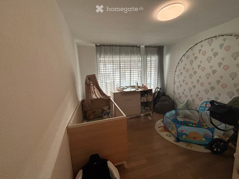 A well-lit children's bedroom with a crib, changing table, playpen, and storage unit. The walls feature a balloon-themed mural, and the room includes a baby stroller and a toy box.