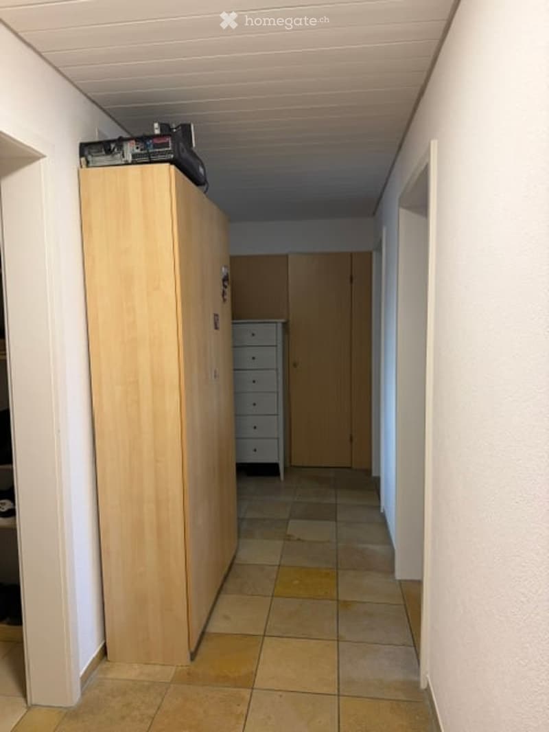 A narrow hallway with tiled flooring, white walls, and a wooden wardrobe on the left side. A small dresser is visible further down the hallway.