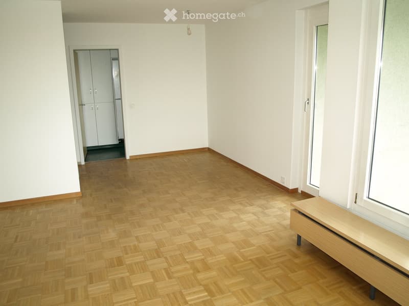 Empty room with wooden parquet flooring, white walls, and a small wooden bench near a window. The room has a doorway leading to a closet area.