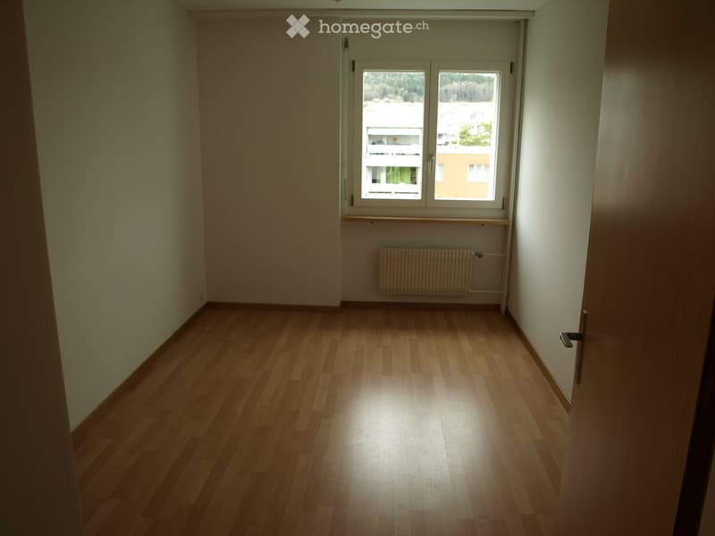 Empty room with wooden flooring, white walls, a window with a view, a radiator, and a door.