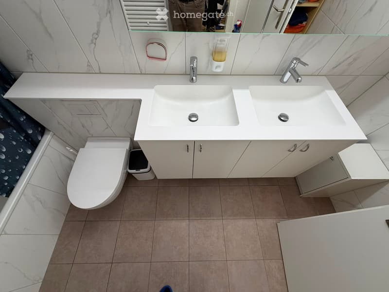 Modern bathroom with white marble tiles, double sink, toilet, and mirrored cabinet.