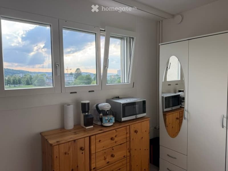 A small, well-lit room with a wooden cabinet holding a coffee maker, microwave, and other small appliances. There is a large mirror on the cabinet and a window with a scenic view.