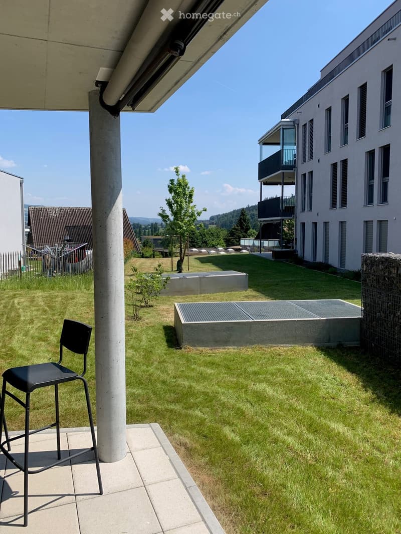 Outdoor area with a grassy lawn, a modern chair, and a view of a contemporary building with balconies and a clear blue sky.