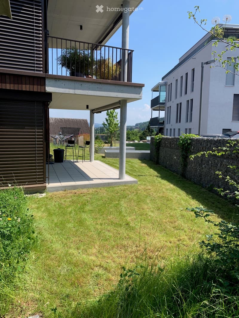 Modern outdoor space with a grassy lawn, a raised concrete platform, a small seating area with two chairs, and a trash bin. The area is bordered by a stone wall and a metal fence, with neighboring buildings visible in the background.