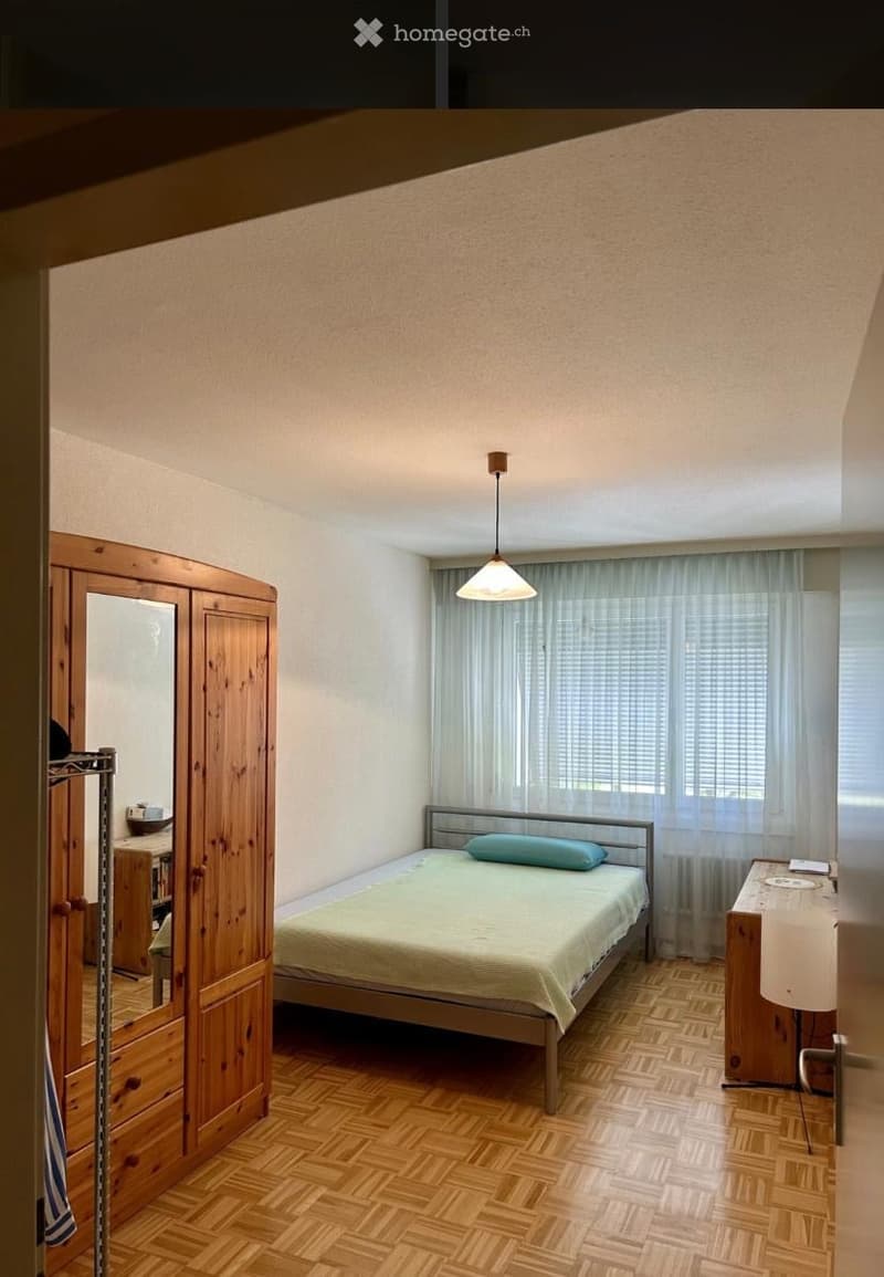 Cozy bedroom with wooden parquet flooring, a single bed with a light green duvet and pillow, a wooden wardrobe with a mirror, a small desk with a lamp, and a window with sheer curtains.