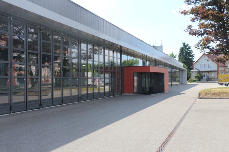 Large building with several rows of glass doors, concrete driveway, house on right, grassy patch