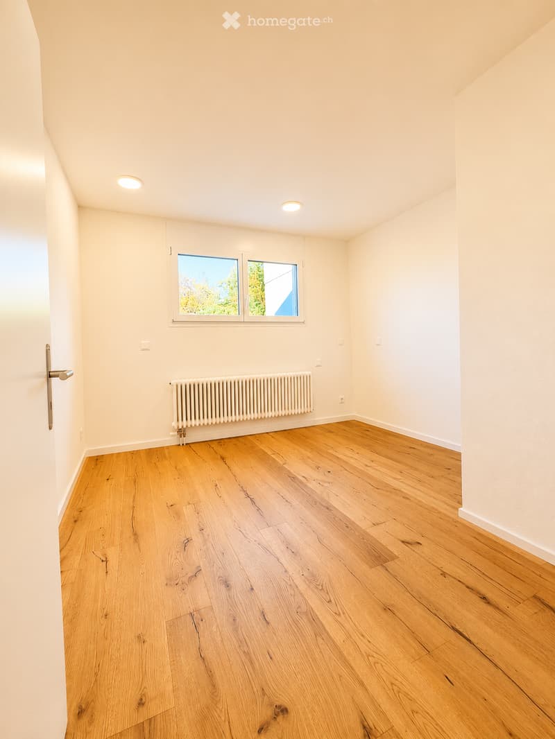 Empty room with wooden flooring, white walls, and a radiator. Features a window with a view of greenery and two ceiling lights.