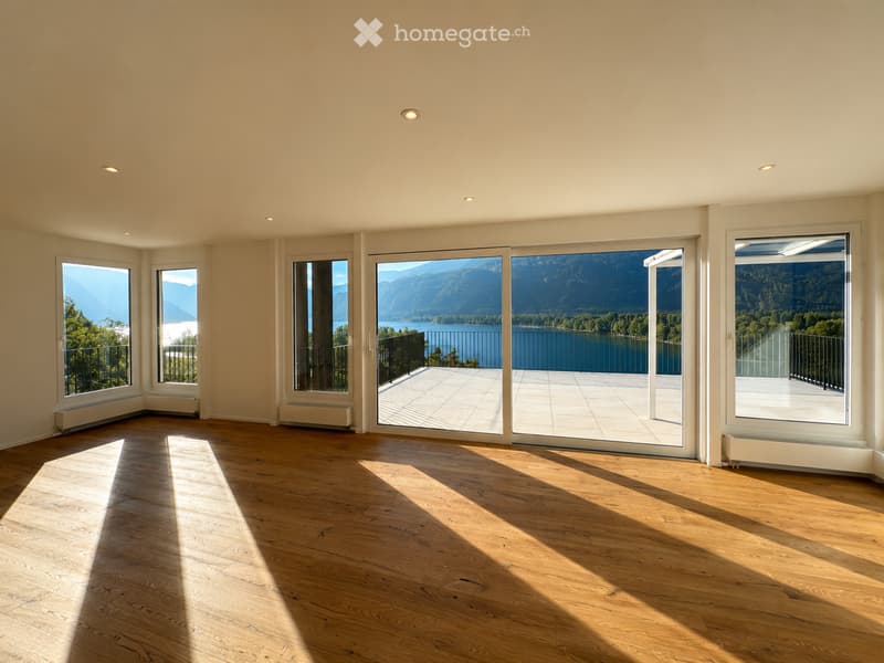 Empty room with wooden flooring, large windows, and a sliding glass door leading to a balcony with a scenic lake and mountain view.