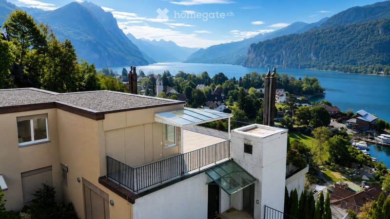 Modern two-story house with a balcony and glass canopy, overlooking a lake and mountain range.