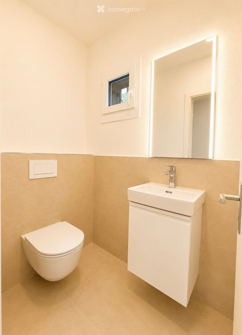 Modern bathroom with beige tiles, white toilet, sink with cabinet, and illuminated mirror above the sink.