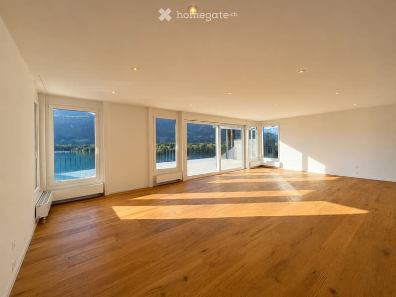 Bright, empty room with wooden flooring and large windows offering a scenic view of a lake and mountains. The space is unfurnished, with natural light flooding in from multiple windows and sliding glass doors.
