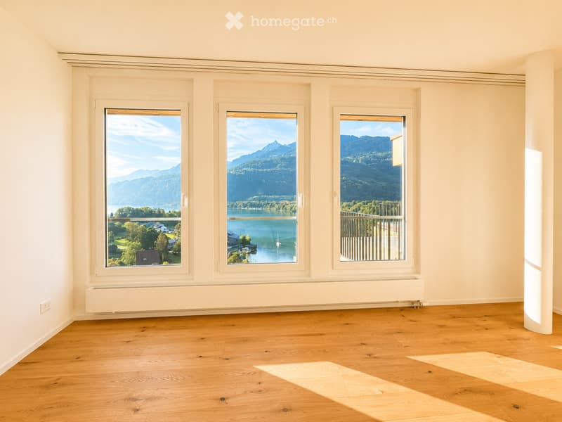 Empty room with wooden flooring, large windows offering scenic mountain and lake views, white walls, and radiators.
