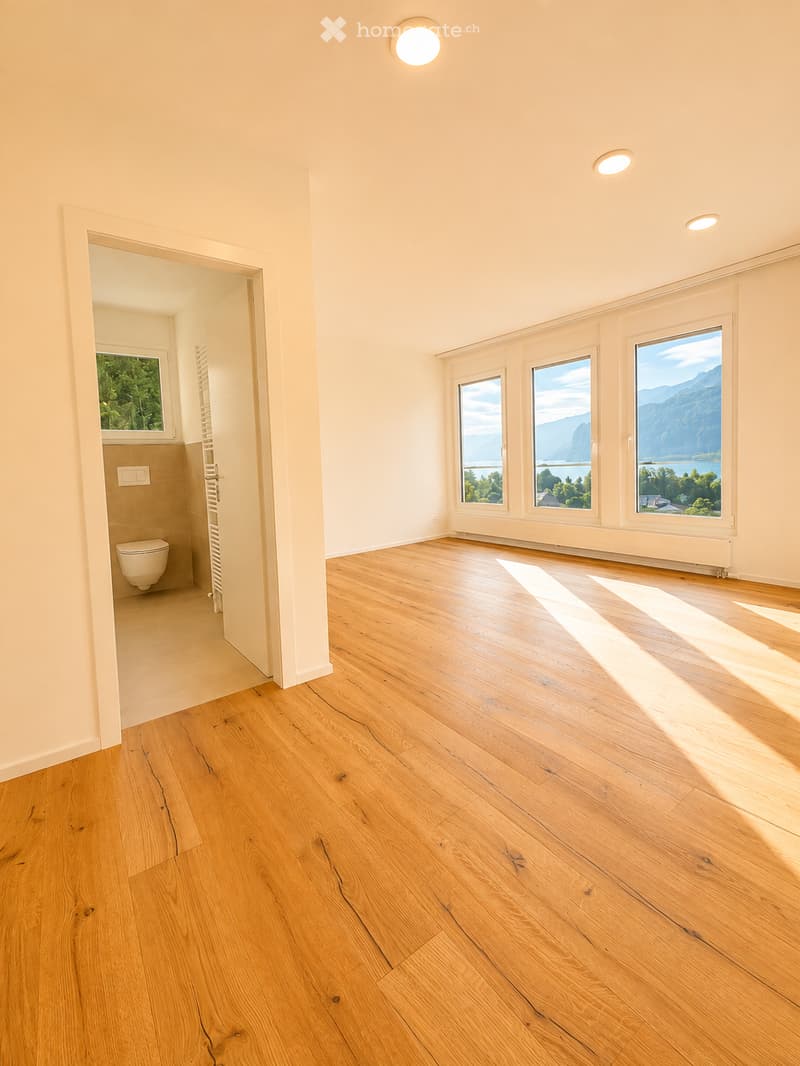 Empty room with wooden flooring, large windows offering scenic views, and a small adjacent bathroom with a toilet.