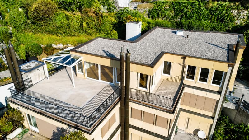 A modern two-story house with a grey tiled roof, multiple balconies, large windows, and a surrounding garden area.
