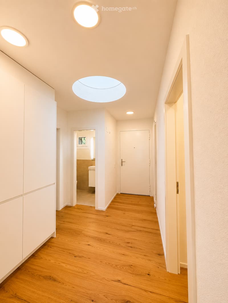 Modern hallway with wooden flooring, white walls, and several round ceiling lights. Doors lead to adjacent rooms, including a visible bathroom.