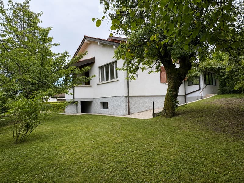 A two-story white house with a red-tiled roof, surrounded by a well-maintained lawn and trees. The house features large windows and a small balcony on the upper floor.