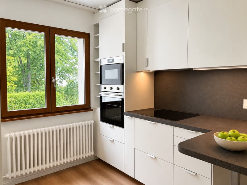 Modern kitchen with white cabinets, dark countertops, built-in oven, and a bowl of green apples on the counter. Large windows offer a view of greenery outside.