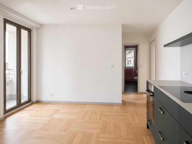 Modern, unfurnished room with light wooden flooring, white walls, and a sliding glass door leading to a balcony. A kitchen area with dark grey cabinets and a built-in oven is visible.