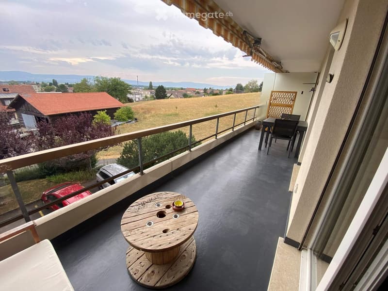 Balcony with a view of a field and distant mountains, featuring a wooden table, a chair, and a small dining set.