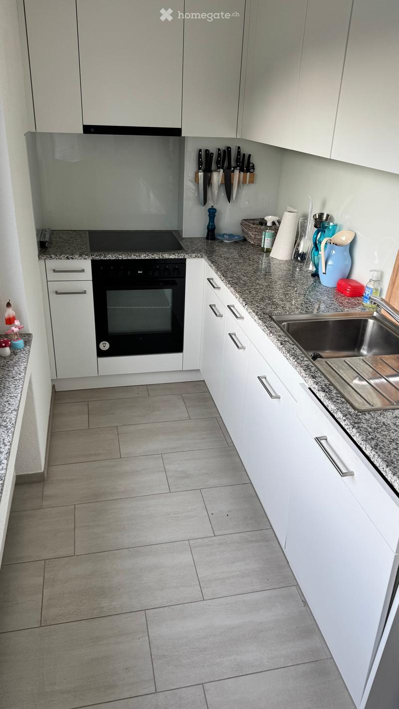 Modern kitchen with white cabinets, granite countertops, and tiled floor. Features an oven, sink, and various small appliances and utensils.