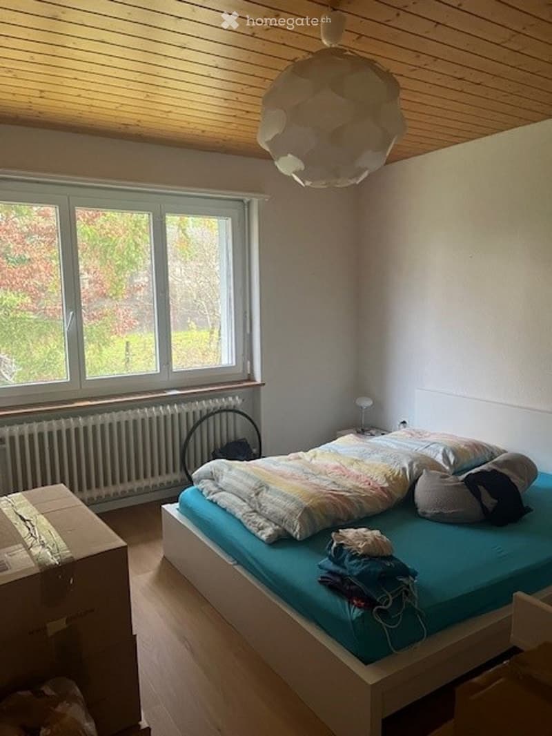 Cozy bedroom with wooden floors and slanted wooden ceiling. Features a single bed with blue mattress, colorful blanket, pillows, and scattered clothing. A window with white frame and radiator below, plus a nightstand with lamp.