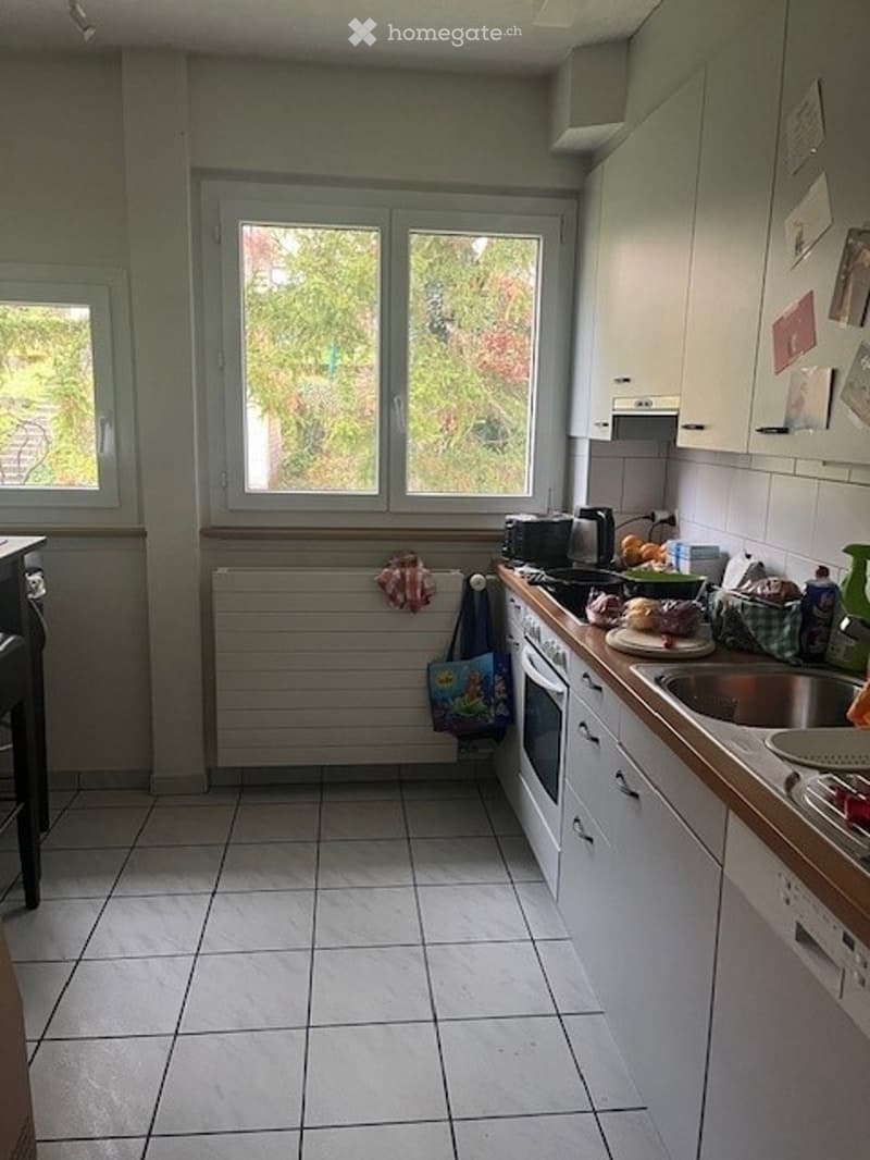 A small kitchen with tiled flooring, white cabinets, and a countertop with various appliances including an oven, microwave, toaster, and sink. There are also some items on the counter such as a basket and a bowl.