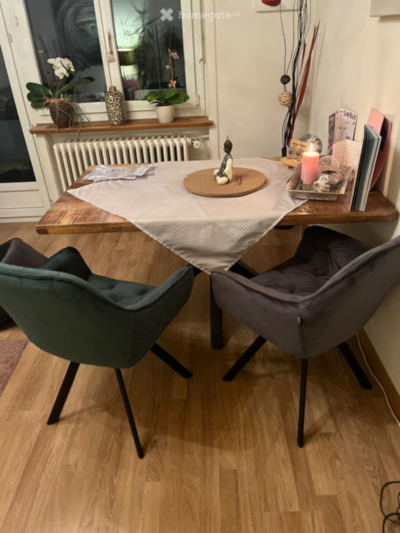 A modern, well-lit room with a wooden table, two chairs, a candle, books, a decorative vase, a small statue, and potted plants on the windowsill.