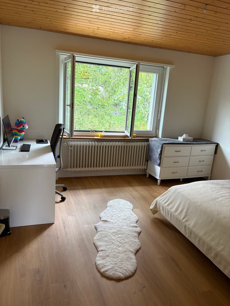 Cozy bedroom with wooden flooring, large window with open shutters, bed with white bedding, desk with computer and chair, drawer unit, and a white shaggy rug.