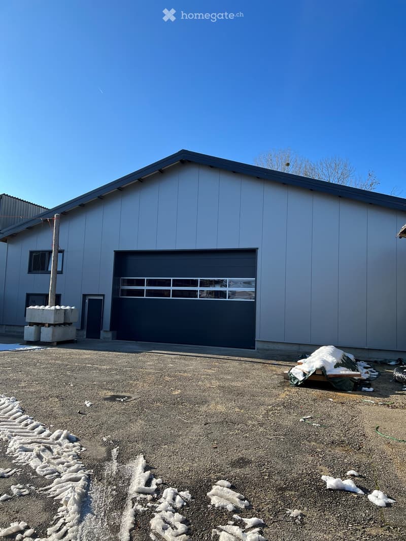 A modern, single-story building with a grey metal exterior, large garage door, and small windows. The surrounding area has gravel ground and scattered snow patches.