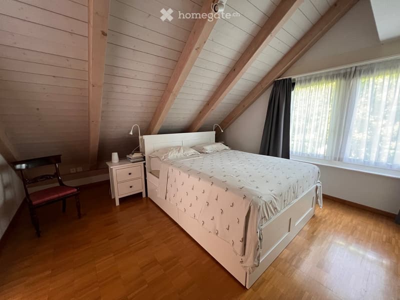 Cozy attic bedroom with wooden slanted ceilings, a double bed with patterned bedding, a nightstand with a lamp, a wooden chair, and a window with dark curtains.