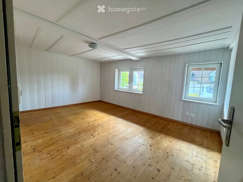 Empty room with wooden flooring, white paneled walls, and two windows offering a view of the outside.