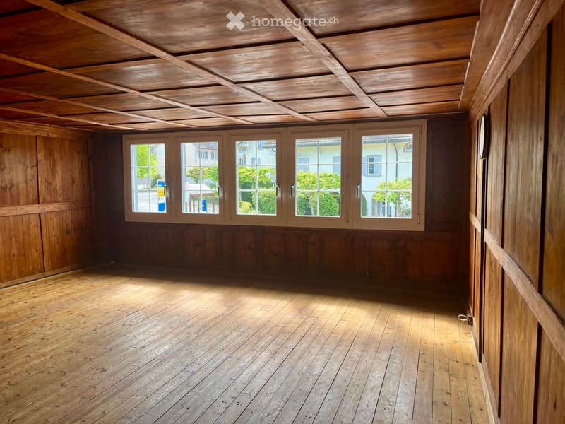 Empty wooden room with exposed wooden ceiling beams, wooden walls, and large windows showing a view of the street and surrounding buildings.