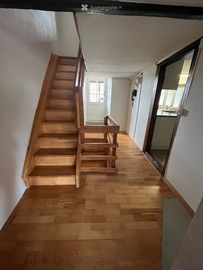 Wooden staircase with a handrail leads up, wooden floor, white walls, small window, and a door leading to another room.