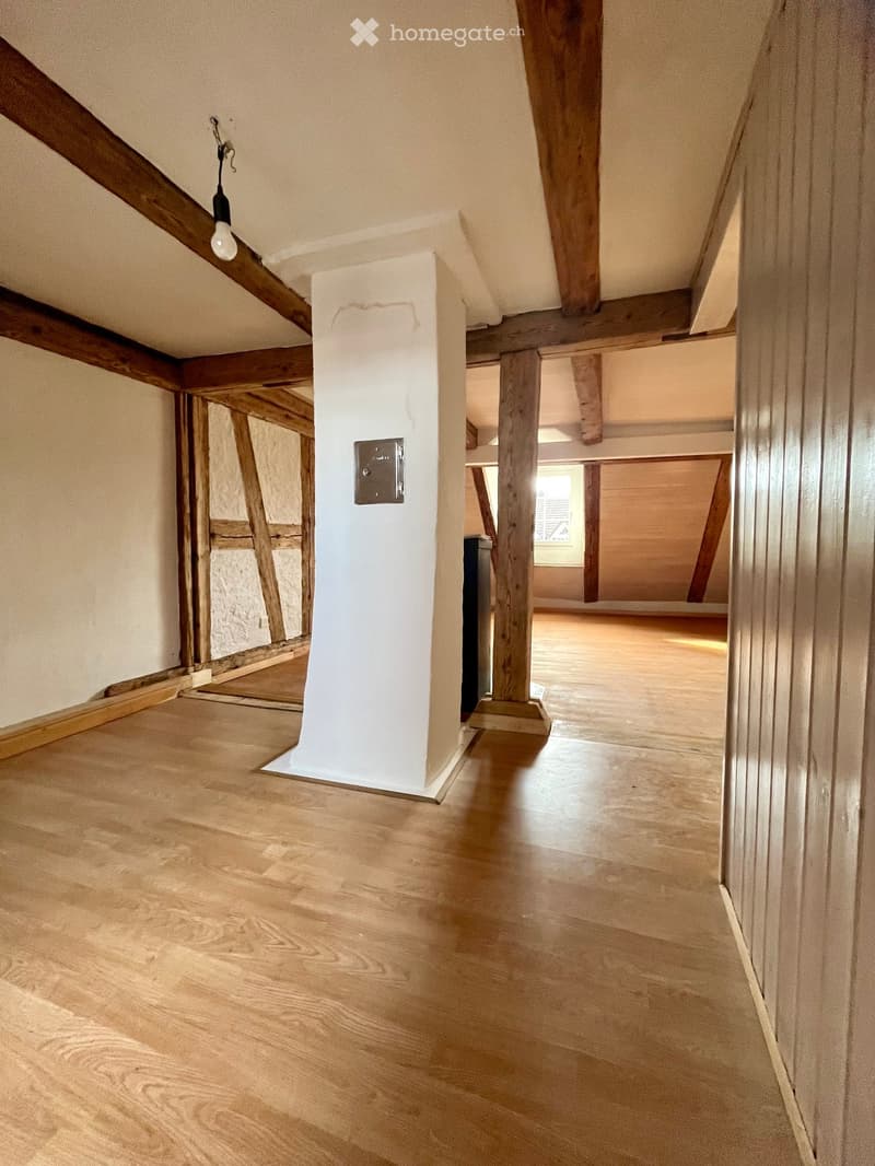 Empty attic space with wooden beams, exposed wooden floor, and a single light fixture hanging from the ceiling.