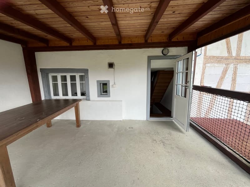 Balcony with wooden table, open wooden ceiling beams, white walls, and a staircase leading down.